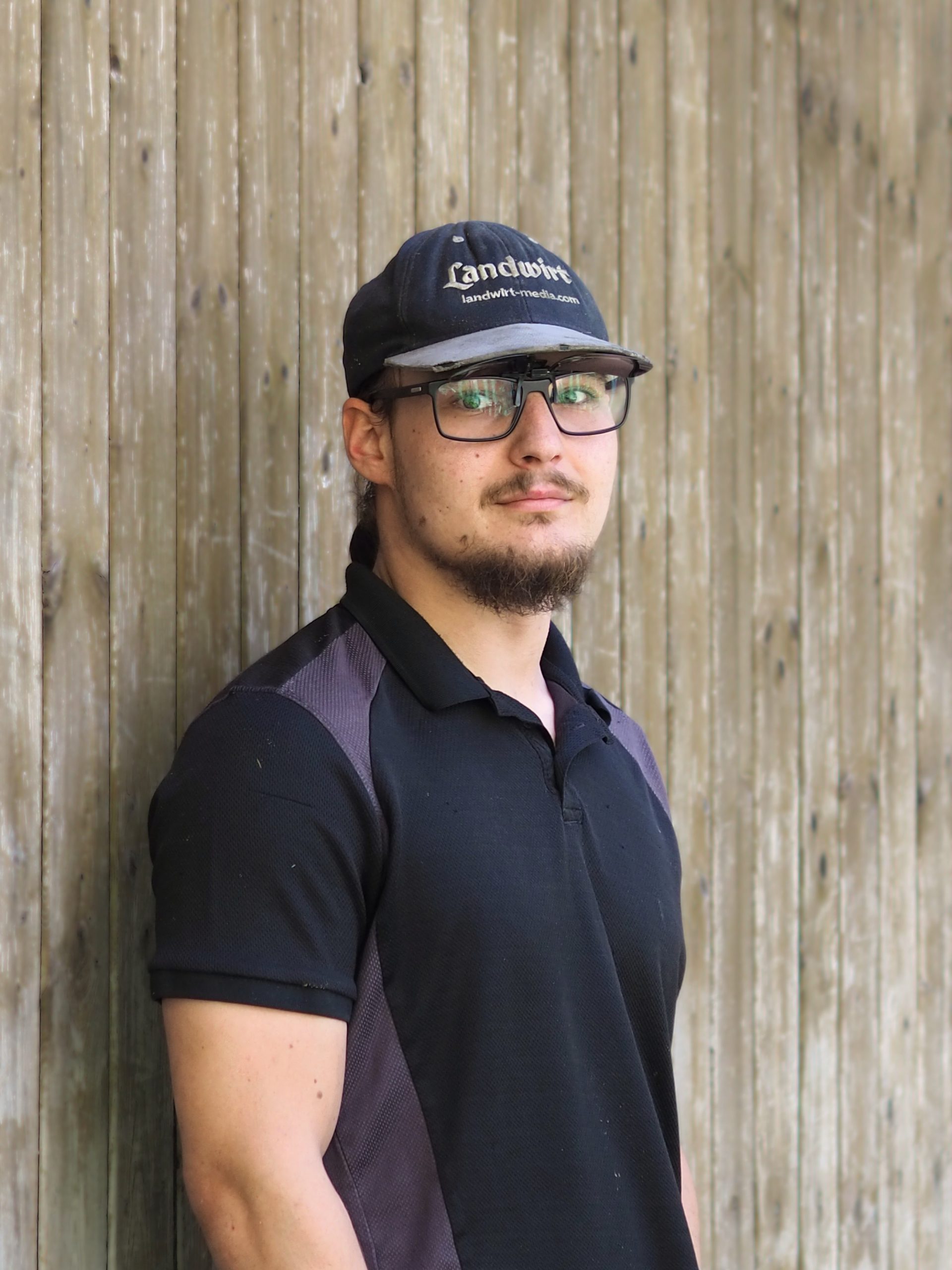 Mann mit Bart, Cap und Brille vor einer Holzwand