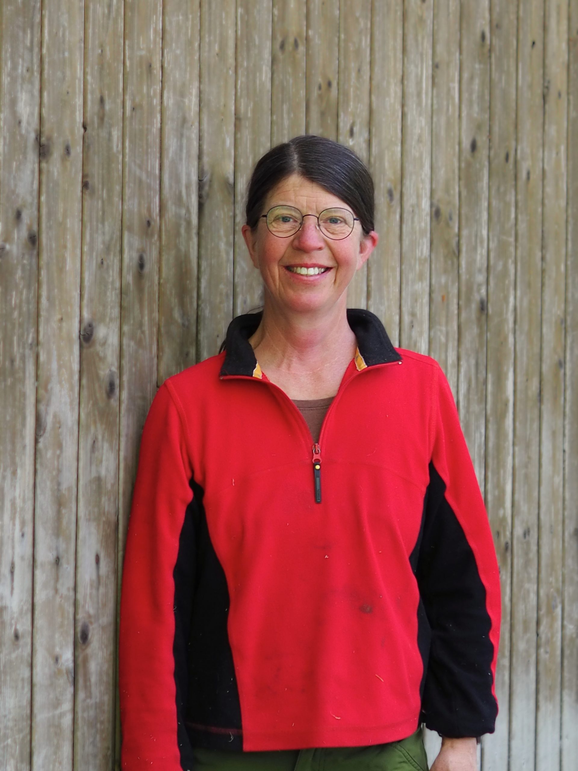 Frau mit Brille, geschlossenen, braunen Haaren und rotem Pullover vor einer Holzwand