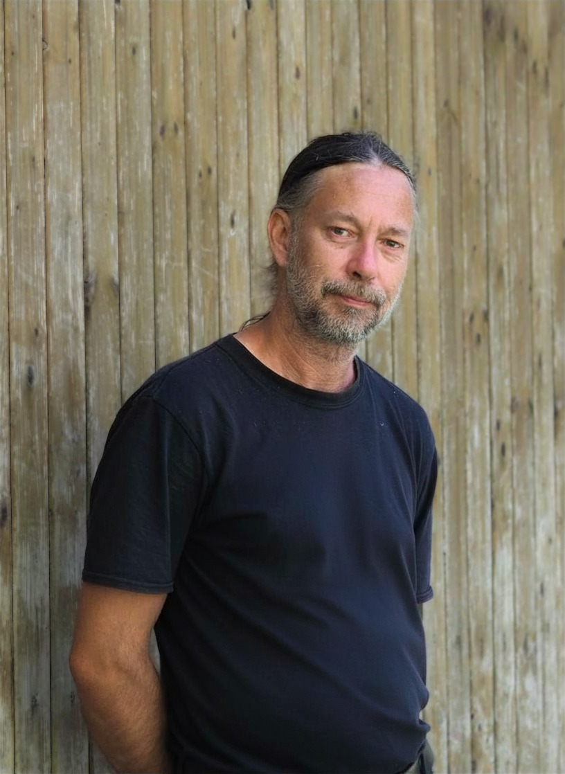Mann mit langen Haaren und schwarzem Shirt vor einer Holzwand