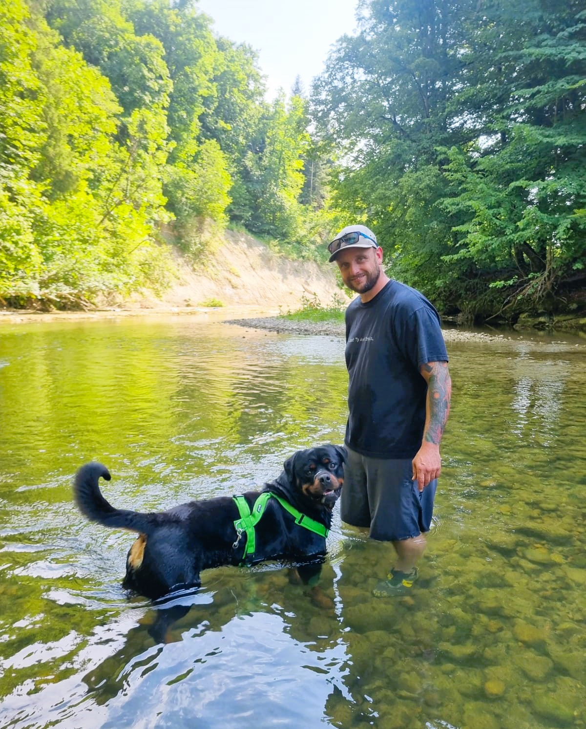 Mann mit Cap und kurzen dunklen Klamotten steht kniehoch in einem Bach mit einem Rottweiler Rüde. Beide genießen das kühle Wasser.