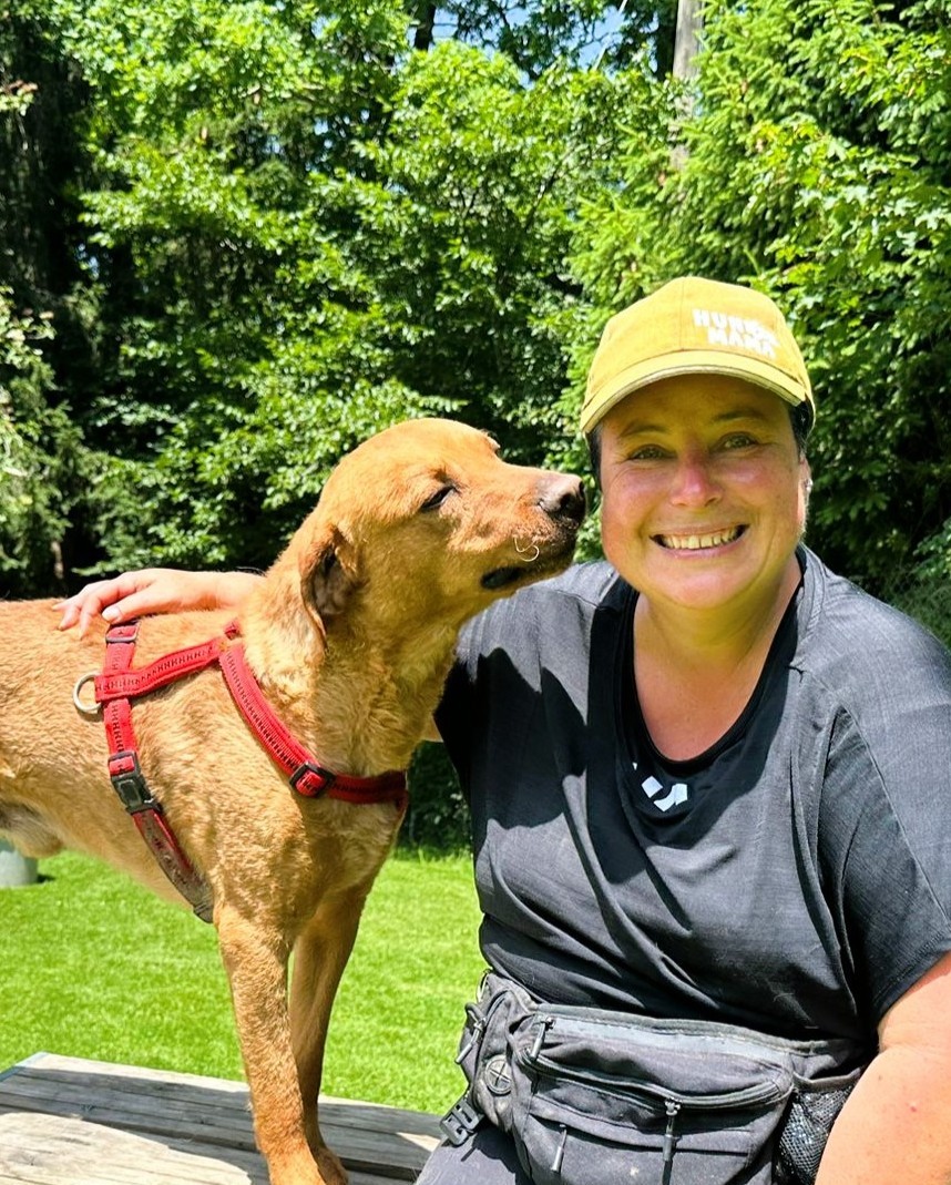Frau mit kurzen Haaren und gelber Cap lacht in die Kamera, währenddessen schnuppert goldbrauner Hund an ihrer Backe.
